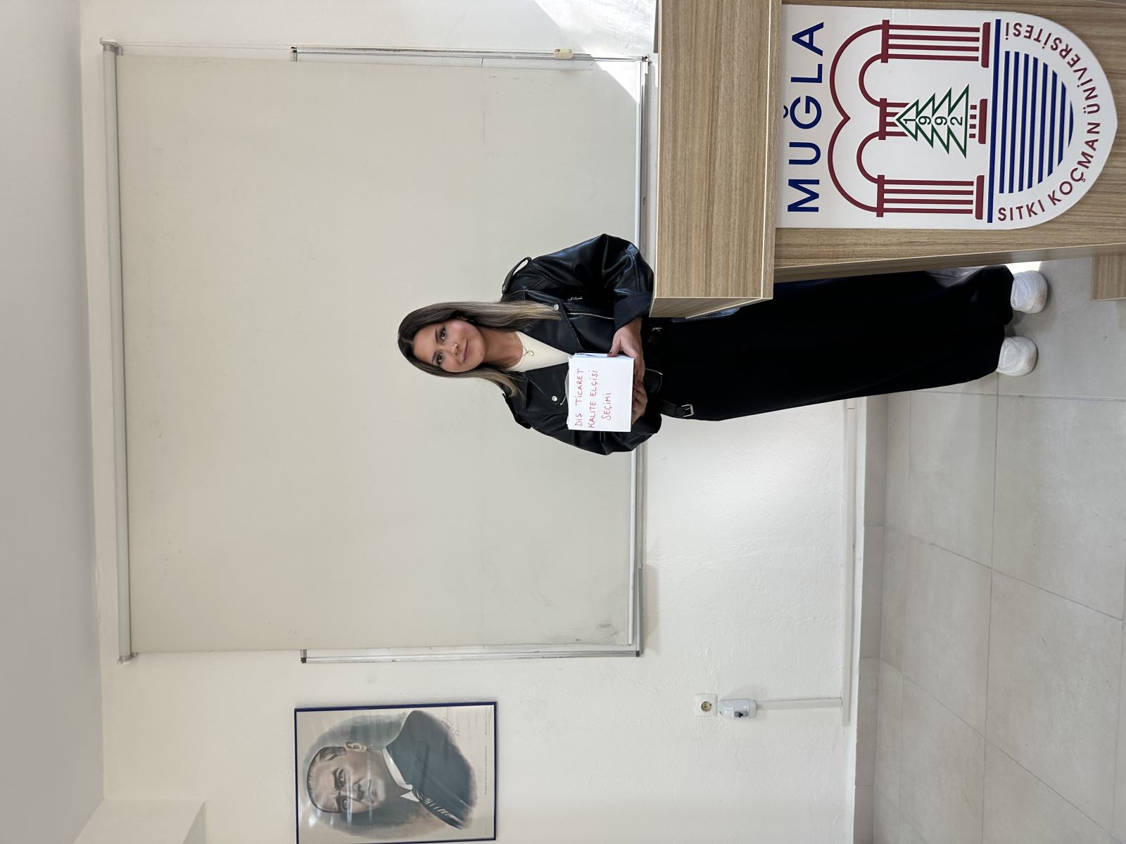 This photo is also taken in the same classroom. A young woman is standing next to the lectern, holding the same box labeled “Foreign Trade Quality Ambassador Election” in red letters. She is wearing a black leather jacket, black pants, and white sneakers. Behind her is a white screen, and to her left is a portrait of Atatürk. The university logo on the lectern reads “Muğla Sıtkı Koçman University.”